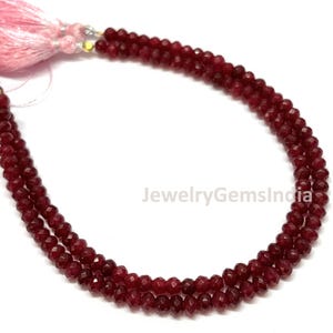 Op de afbeelding: Een streng kleine, gefacetteerde, dieprode edelsteenkralen in een gebogen vorm. De kralen zijn geregen aan een dunne draad en afgewerkt met lichtroze decoratieve kwastjes aan de uiteinden. De tekst JewelryGemsIndia is zichtbaar in het midden.