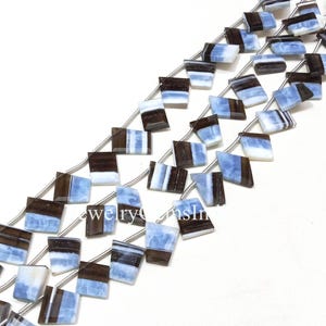 May include: Strands of square-cut beads in shades of blue, white, and brown. The beads are strung on a silver-colored wire, creating a pattern of alternating colors. The beads are arranged diagonally across the frame.
