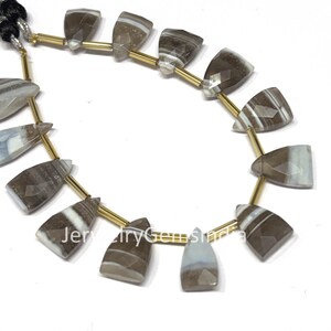 Könnte beinhalten: Eine Kette aus Edelsteinperlen. Jede Perle hat eine trapezförmige Form mit Facetten und weist Brauntöne, Weiß und Grau auf. Die Perlen sind auf goldfarbenem Draht aufgefädelt, mit einer schwarzen Quaste oben. Die Perlen sind etwa 2.5 cm lang.