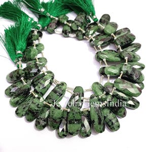 Peut inclure: Collier de perles de pierres précieuses vertes et noires, taillées à facettes. Les perles sont enfilées sur un fil argenté et agrémentées de pompons verts. Les perles ovales ont une finition polie.