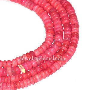 May include: Close-up of strands of pink and red gemstone beads. The beads are small, round, and uniform in size, with a slightly flattened shape. The text "JewelryGemsIndia" is visible.