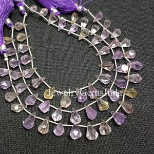 May include: Three strands of gemstone beads in shades of purple, yellow, and clear. The teardrop-shaped beads are strung on silver wire. Purple tassels are at the top. The beads are displayed on a dark grey surface.