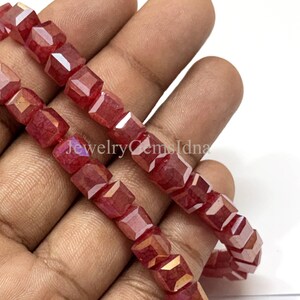 Könnte beinhalten: Stränge facettierter, roter Perlen mit einer geometrischen, würfelförmigen Form. Die Perlen haben eine reflektierende, irisierende Qualität und werden in der Hand einer Person gehalten. Der Text "jewelrygemsidna" ist sichtbar.