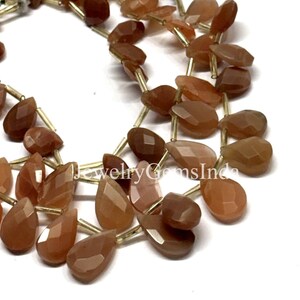 May include: Close-up of strands of faceted, teardrop-shaped gemstones in shades of brown and orange. The gemstones are strung together with gold-colored wire, creating a necklace or bracelet. The gemstones have a polished, reflective surface.