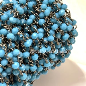 Peut inclure: Gros plan sur un fil de perles bleues claires à facettes enfilées sur une chaîne en métal foncé. Les perles sont d'un bleu turquoise vif et les maillons de la chaîne sont visibles. Le texte "Jewelry Gems India" est en bas à droite.