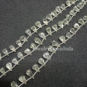 May include: Close-up of several strands of clear, faceted teardrop-shaped beads strung on a thin, silver-coloured wire. The beads are arranged in rows against a dark grey textured background. The text "JewelryGemsInda" is visible.