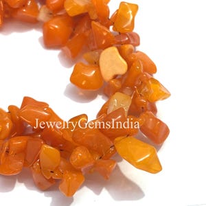 May include: Close-up of a collection of polished, orange-colored gemstone chips. The chips are irregularly shaped and vary in size, with a glossy finish. The text "JewelryGemsIndia" is visible in the image.