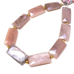 Könnte beinhalten: Eine Kette aus rechteckigen, facettierten Perlen in Rosa- und Grautönen. Jede Perle wird durch eine kleine goldene Perle getrennt. Die Perlen sind aufgefädelt, mit einer Quaste am Ende.