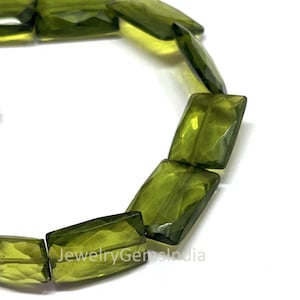 Könnte beinhalten: Nahaufnahme eines Strangs olivgrüner, rechteckiger Edelsteinperlen. Die Perlen haben eine facettierte Oberfläche, die das Licht einfängt. Der Hintergrund ist weiß. Der Text "JewelryGemsIndia" ist unten rechts sichtbar.