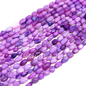 Könnte beinhalten: Nahaufnahme von Strängen aus lila Edelsteinperlen. Die Perlen sind oval geformt und variieren in den Lilatönen, von hellem Lavendel bis zu tiefem Violett. Die Perlen sind glatt und poliert, mit leichtem Glanz.