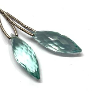 Könnte beinhalten: Zwei facettierte, hellblaue Edelsteine im Marquise-Schliff, die an dünnen, flexiblen, silberfarbenen Metallspiral-Ketten hängen. Die Steine haben eine spitze, blattartige Form mit einer reflektierenden Oberfläche, die ein klares, durchscheinendes Aussehen erzeugt.