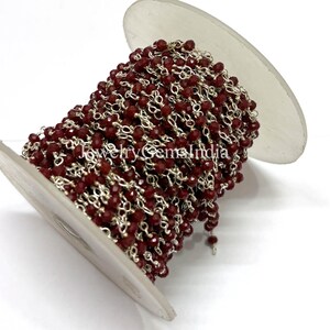Könnte beinhalten: Eine Spule mit Perlenkette mit kleinen, facettierten, dunkelroten Perlen, die auf silberfarbenen Gliedern aufgereiht sind. Die Kette ist eng um eine weiße Spule gewickelt. Der Text "Jewelry Gems India" ist auf der Spule zu sehen.