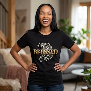 May include: A black short-sleeved t-shirt featuring a large, ornate, cream-coloured letter B with the word BLESSED printed in a metallic gold, serif font across the centre of the letter. The design is positioned centrally on the chest.