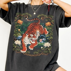 K&ouml;nnte beinhalten: Dunkelgraues T-Shirt mit einer lebendigen Illustration eines Tigers in einer nat&uuml;rlichen Umgebung. Der Tiger ist mit orangefarbenen und schwarzen Streifen dargestellt, umgeben von Blumen und Bl&auml;ttern. Das Shirt hat kurze &Auml;rmel und eine lockere Passform.