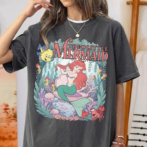 May include: A charcoal gray short-sleeve t-shirt featuring a graphic print of The Little Mermaid. The design displays Ariel with red hair, Flounder, and Sebastian surrounded by underwater coral and seaweed. The text reads The Little Mermaid.