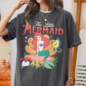 May include: A charcoal gray t-shirt featuring a graphic print of The Little Mermaid with Ariel sitting in a large seashell, accompanied by Flounder and Sebastian. The text The Little Mermaid is printed in red above the character illustration.
