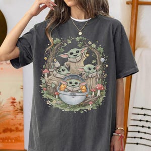 May include: A charcoal gray short-sleeve t-shirt featuring a graphic print of four small green creatures with large ears. The illustration shows them in a forest setting with mushrooms, foliage, and one creature sitting inside a metallic pod.