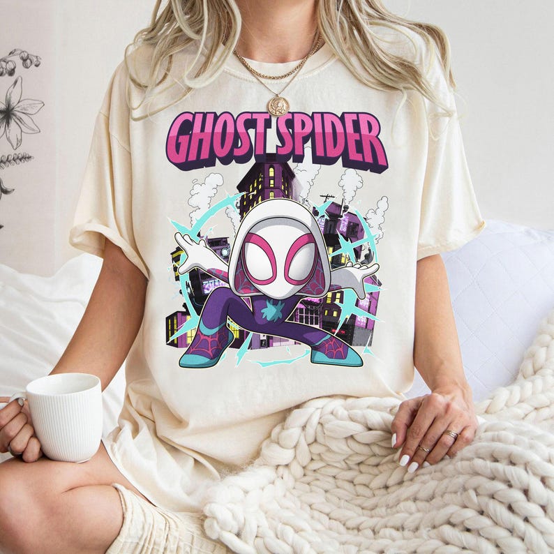 May include: Cream-colored t-shirt featuring the words "GHOST SPIDER" in pink and purple. The graphic depicts a cartoon version of Ghost-Spider in a dynamic pose, set against a cityscape background. The character is wearing a white and pink mask and a purple and blue suit.