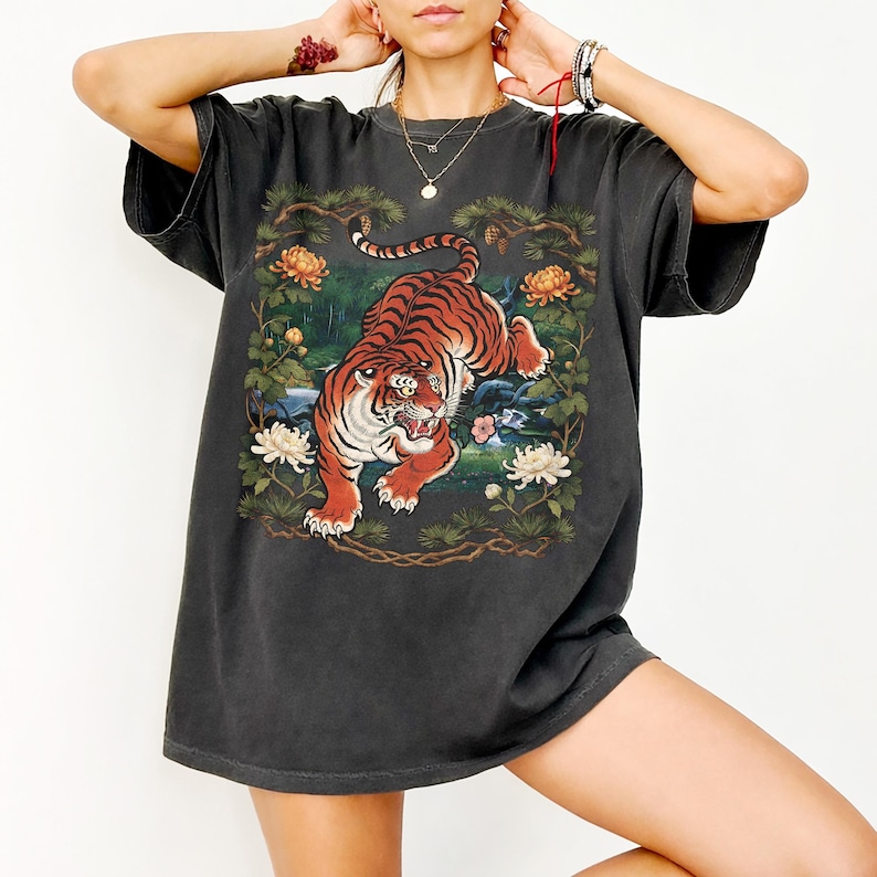 K&ouml;nnte beinhalten: Dunkelgraues T-Shirt mit einer lebendigen Illustration eines Tigers in einer nat&uuml;rlichen Umgebung. Der Tiger ist mit orangefarbenen und schwarzen Streifen dargestellt, umgeben von Blumen und Bl&auml;ttern. Das Shirt hat kurze &Auml;rmel und eine lockere Passform.
