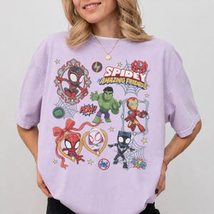 以下が含まれることがあります： ハルク、アイアンマン、スパイダーマンなどの「スパイディとすごいなかまたち」のキャラクターが描かれた、ライトパープルの半袖Tシャツ。デザインには装飾的なフレーム、クモの巣、そして「Spidey Amazing Friends」というテキストが含まれています。