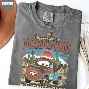 May include: A grey Comfort Colors t-shirt with a graphic of Mater from the film Cars, and the text "Happier than a Tornado." The design includes a desert landscape with cacti and a setting sun. A gold necklace is draped across the shirt.