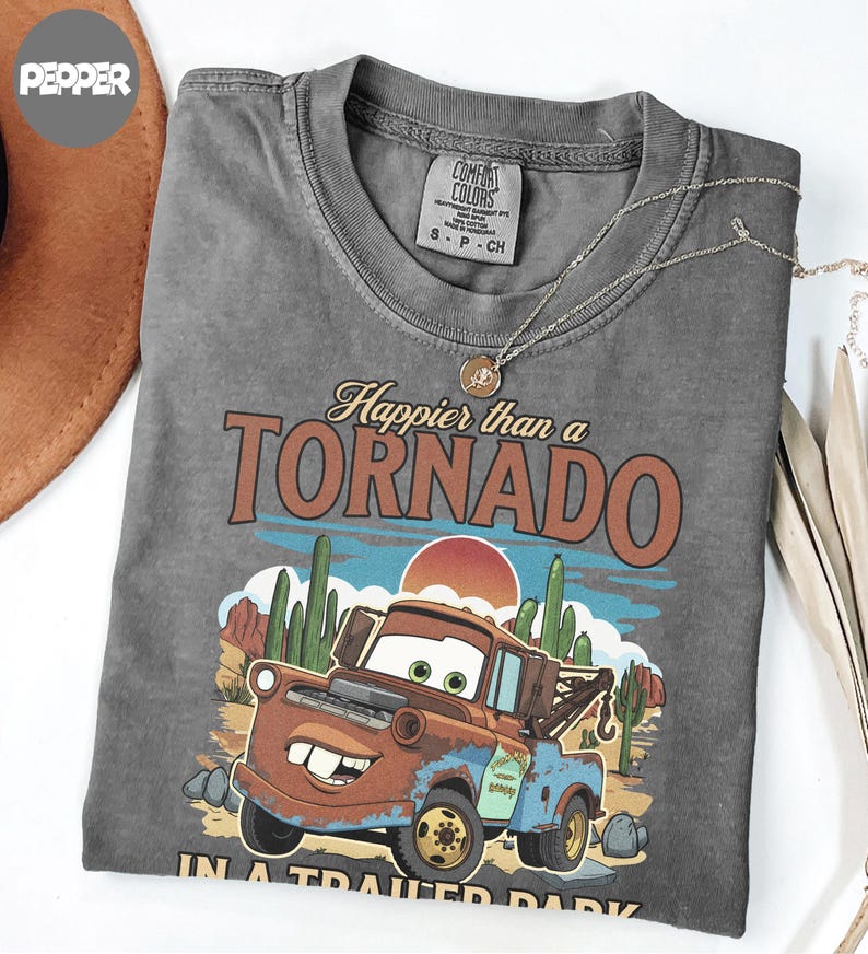 May include: A grey Comfort Colors t-shirt with a graphic of Mater from the film Cars, and the text "Happier than a Tornado." The design includes a desert landscape with cacti and a setting sun. A gold necklace is draped across the shirt.