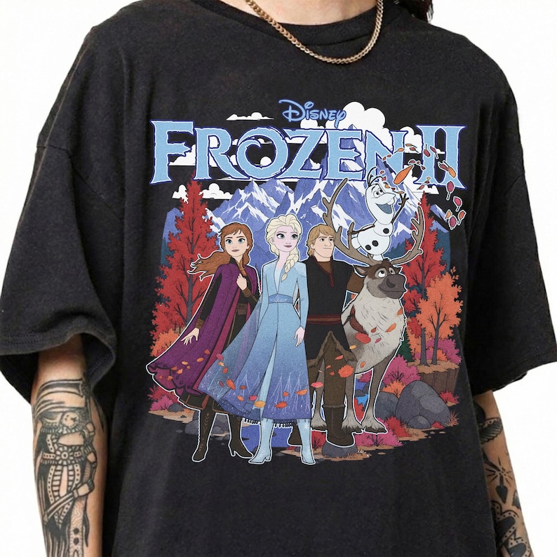 May include: A black short-sleeved t-shirt featuring a graphic print of the Disney Frozen II logo above characters Anna, Elsa, Kristoff, Sven, and Olaf. The background depicts a colorful autumn forest with mountains under a cloudy sky.