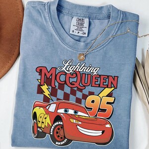 May include: A light blue short-sleeve t-shirt featuring a graphic of the red race car Lightning McQueen with the number 95, a checkered flag background, and yellow lightning bolts. The text reads Lightning MCQUEEN 95. The shirt has a Comfort Colors label.