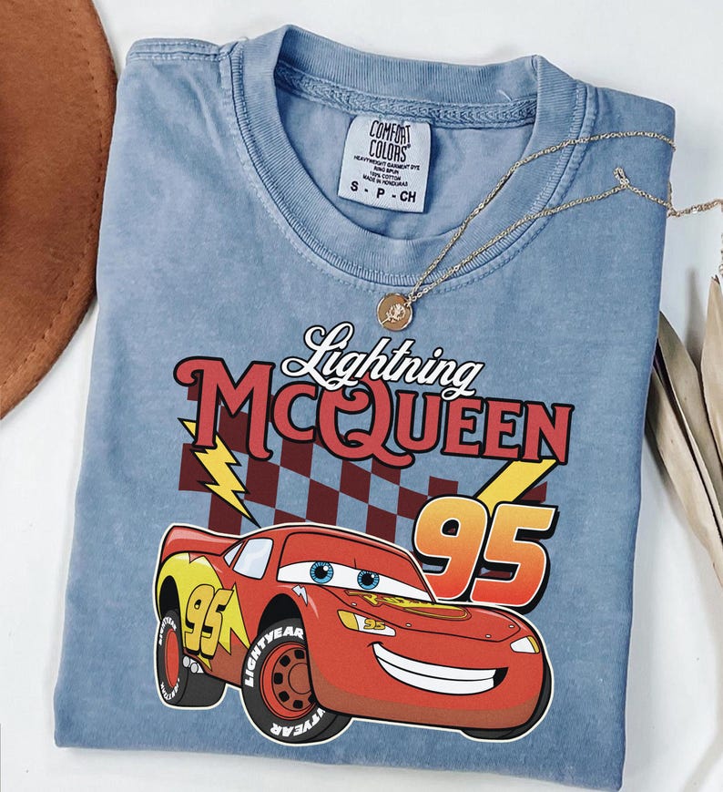 May include: A light blue short-sleeve t-shirt featuring a graphic of the red race car Lightning McQueen with the number 95, a checkered flag background, and yellow lightning bolts. The text reads Lightning MCQUEEN 95. The shirt has a Comfort Colors label.