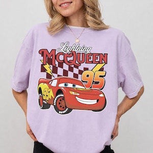 May include: A light purple short-sleeve t-shirt featuring a graphic of the red race car Lightning McQueen with the number 95, a checkered pattern, and the text Lightning MCQUEEN 95 printed in a bold, stylized font across the chest area.