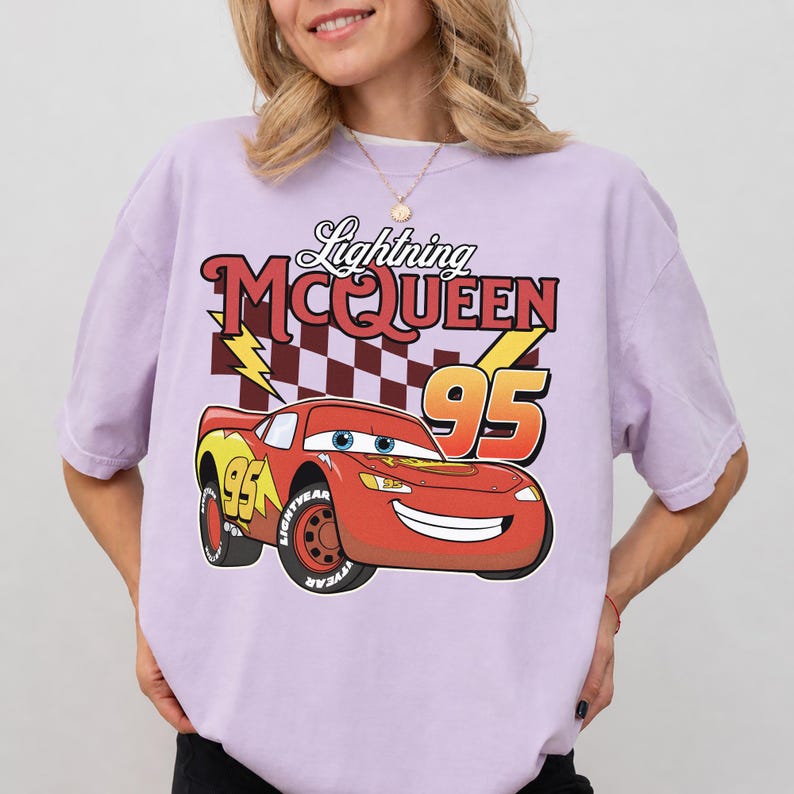 May include: A light purple short-sleeve t-shirt featuring a graphic of the red race car Lightning McQueen with the number 95, a checkered pattern, and the text Lightning MCQUEEN 95 printed in a bold, stylized font across the chest area.