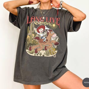 Puede incluir: Una camiseta de manga corta gris carb&oacute;n con un estampado gr&aacute;fico de una vaquera montando un caballo con un lazo. El texto "LONG LIVE cowgirls" est&aacute; impreso sobre la ilustraci&oacute;n, que incluye cactus y un sol. La camiseta tiene un corte holgado.