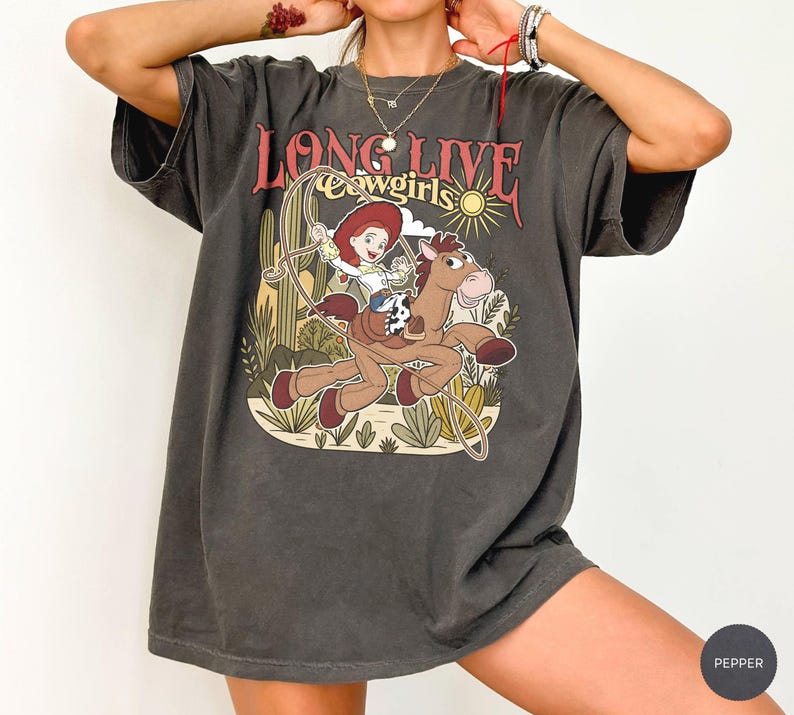 Puede incluir: Una camiseta de manga corta gris carb&oacute;n con un estampado gr&aacute;fico de una vaquera montando un caballo con un lazo. El texto "LONG LIVE cowgirls" est&aacute; impreso sobre la ilustraci&oacute;n, que incluye cactus y un sol. La camiseta tiene un corte holgado.