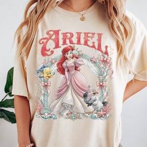 May include: A cream-colored t-shirt featuring a graphic print of Ariel in a pink gown, surrounded by an ornate archway with sea-themed elements including a yellow fish, a seagull, shells, and pearls. The word ARIEL is printed in red above.