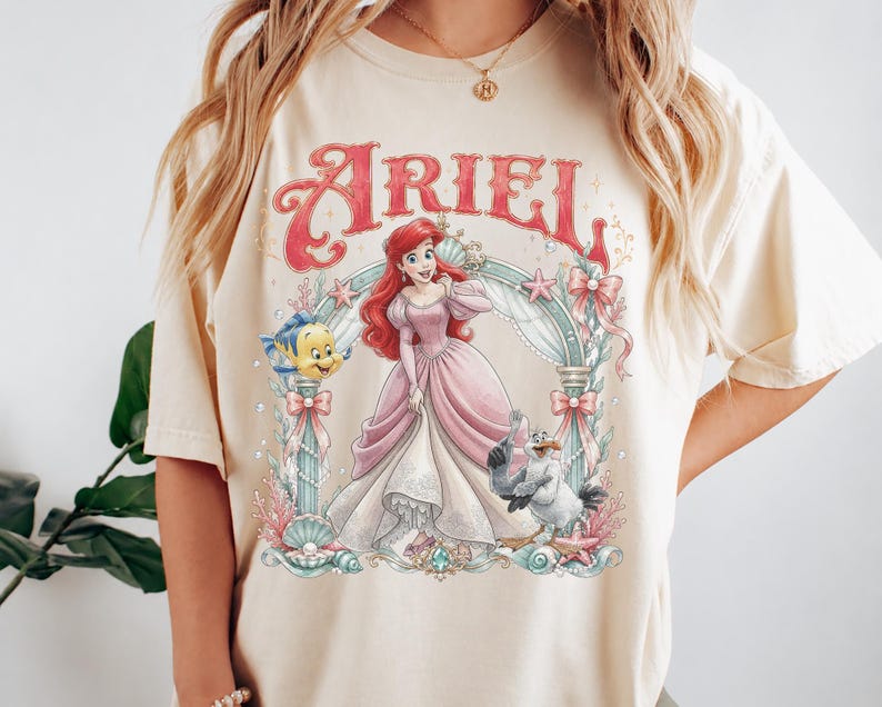 May include: A cream-colored t-shirt featuring a graphic print of Ariel in a pink gown, surrounded by an ornate archway with sea-themed elements including a yellow fish, a seagull, shells, and pearls. The word ARIEL is printed in red above.
