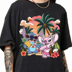 May include: A black short-sleeve t-shirt featuring a graphic print of two blue and pink alien characters on a beach. The blue character wears red sunglasses and a floral lei, holding a pineapple. The pink character wears a bikini top.