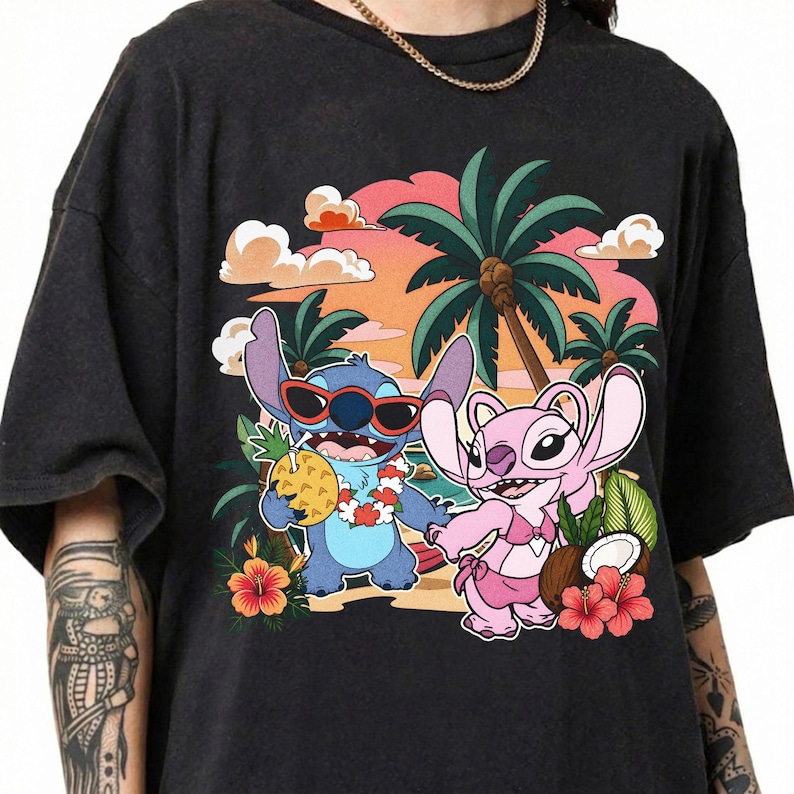 May include: A black short-sleeve t-shirt featuring a graphic print of two blue and pink alien characters on a beach. The blue character wears red sunglasses and a floral lei, holding a pineapple. The pink character wears a bikini top.
