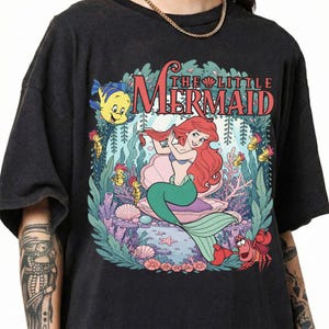 May include: A black short-sleeve t-shirt featuring a graphic print of Ariel from The Little Mermaid sitting in a shell, surrounded by Flounder, Sebastian, and sea life. The text THE LITTLE MERMAID is printed in red above the character illustration.