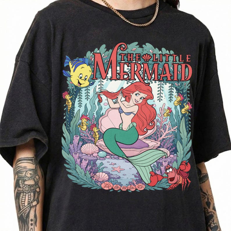 May include: A black short-sleeve t-shirt featuring a graphic print of Ariel from The Little Mermaid sitting in a shell, surrounded by Flounder, Sebastian, and sea life. The text THE LITTLE MERMAID is printed in red above the character illustration.
