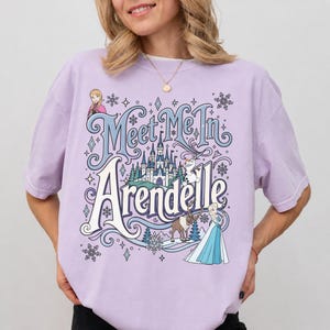 K&ouml;nnte beinhalten: Ein hellviolettes Kurzarm-T-Shirt mit einem Grafikdruck und dem Text Meet Me In Arendelle. Das Design zeigt Illustrationen eines Schlosses, Schneeflocken, Anna, Elsa, Olaf und Sven in den Farben Blau, Wei&szlig; und Braun.