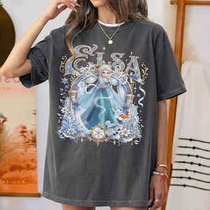 May include: A charcoal gray short-sleeve t-shirt featuring a graphic print of the character Elsa and Olaf from Frozen. The design includes the text Elsa in a decorative font, surrounded by intricate blue and gold floral and snowflake patterns.