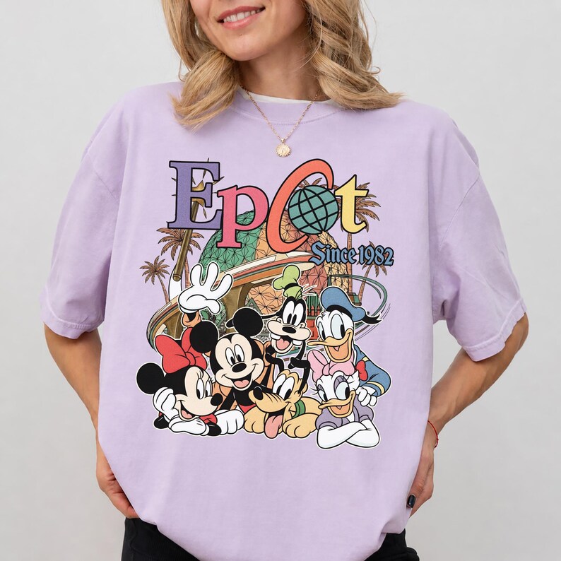 May include: A light purple t-shirt featuring a colorful graphic of Disney characters, including Mickey Mouse, Minnie Mouse, Donald Duck, Goofy, and Pluto. The graphic also includes the text "Epcot Since 1982" in a retro style.