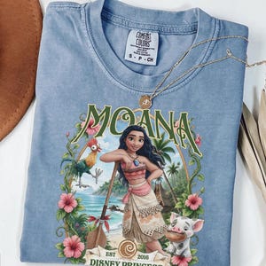 May include: A blue short-sleeve t-shirt featuring a graphic print of the character Moana, a rooster, and a piglet. The design includes the text "MOANA" and "EST 2016 DISNEY PRINCESS" framed by tropical flowers and greenery. The shirt has a Comfort Colors label.