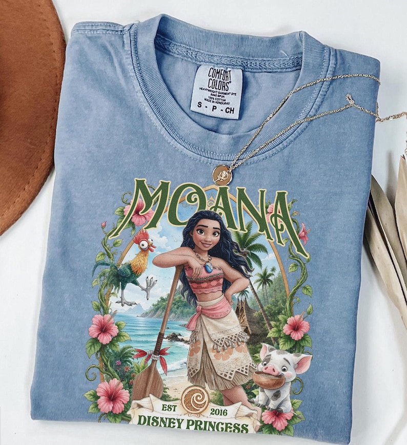 May include: A blue short-sleeve t-shirt featuring a graphic print of the character Moana, a rooster, and a piglet. The design includes the text "MOANA" and "EST 2016 DISNEY PRINCESS" framed by tropical flowers and greenery. The shirt has a Comfort Colors label.