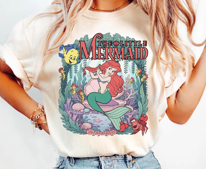 May include: A cream-colored t-shirt featuring a graphic print of The Little Mermaid. The design shows Ariel with red hair, Flounder, and Sebastian surrounded by underwater plants, coral, and seashells. The text THE LITTLE MERMAID is printed above.