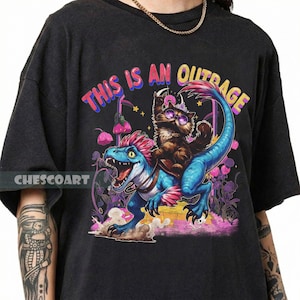 May include: Dark gray t-shirt with a colorful graphic. The graphic features a blue dinosaur with a pink mohawk and a cat wearing sunglasses and a crown, with the text "THIS IS AN OUTRAGE".