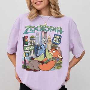 May include: A light purple t-shirt featuring a graphic print of the characters Judy Hopps and Nick Wilde from Zootopia. The design includes the text Zootopia and Bunny Best Friend, set against a colorful city skyline with a traffic light and trees.