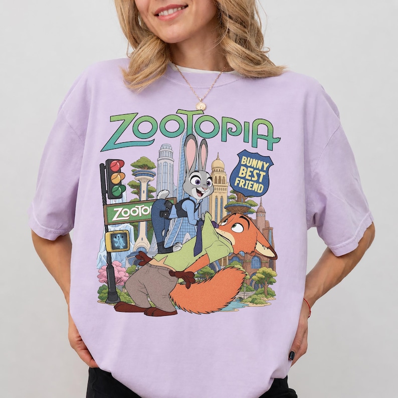 May include: A light purple t-shirt featuring a graphic print of the characters Judy Hopps and Nick Wilde from Zootopia. The design includes the text Zootopia and Bunny Best Friend, set against a colorful city skyline with a traffic light and trees.