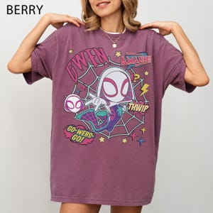May include: A berry-colored short-sleeve t-shirt featuring a graphic of the character Gwen Stacy in a spiderweb design. The print includes the text GWEN, AMAZING, THWIP, and GO-WEBS-GO, along with yellow stars and lightning bolt accents on the front.