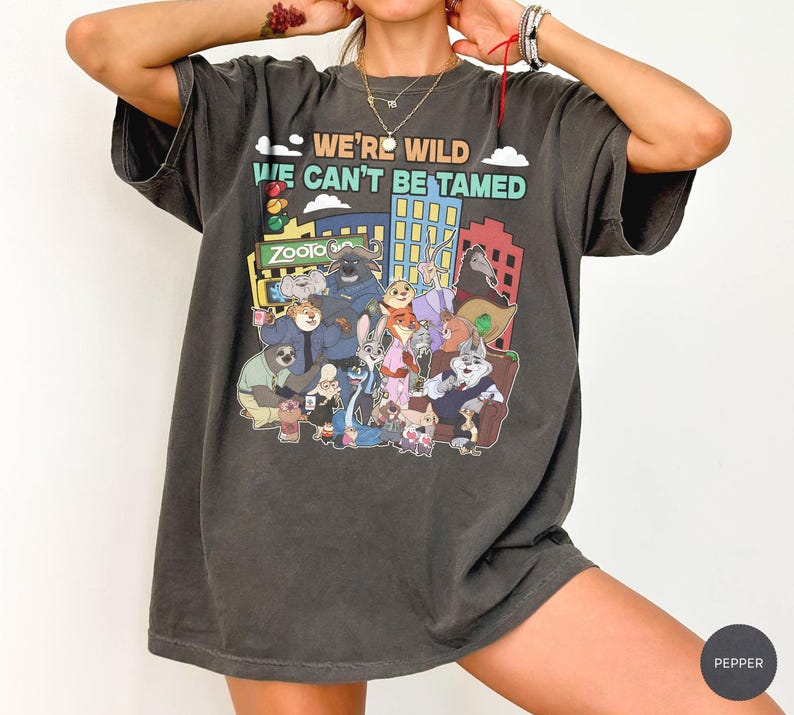 May include: Dark gray t-shirt featuring a colorful graphic of animal characters and the text "WE'RE WILD WE CAN'T BE TAMED". The graphic includes the word "Zootopia". The shirt has short sleeves and a relaxed fit.